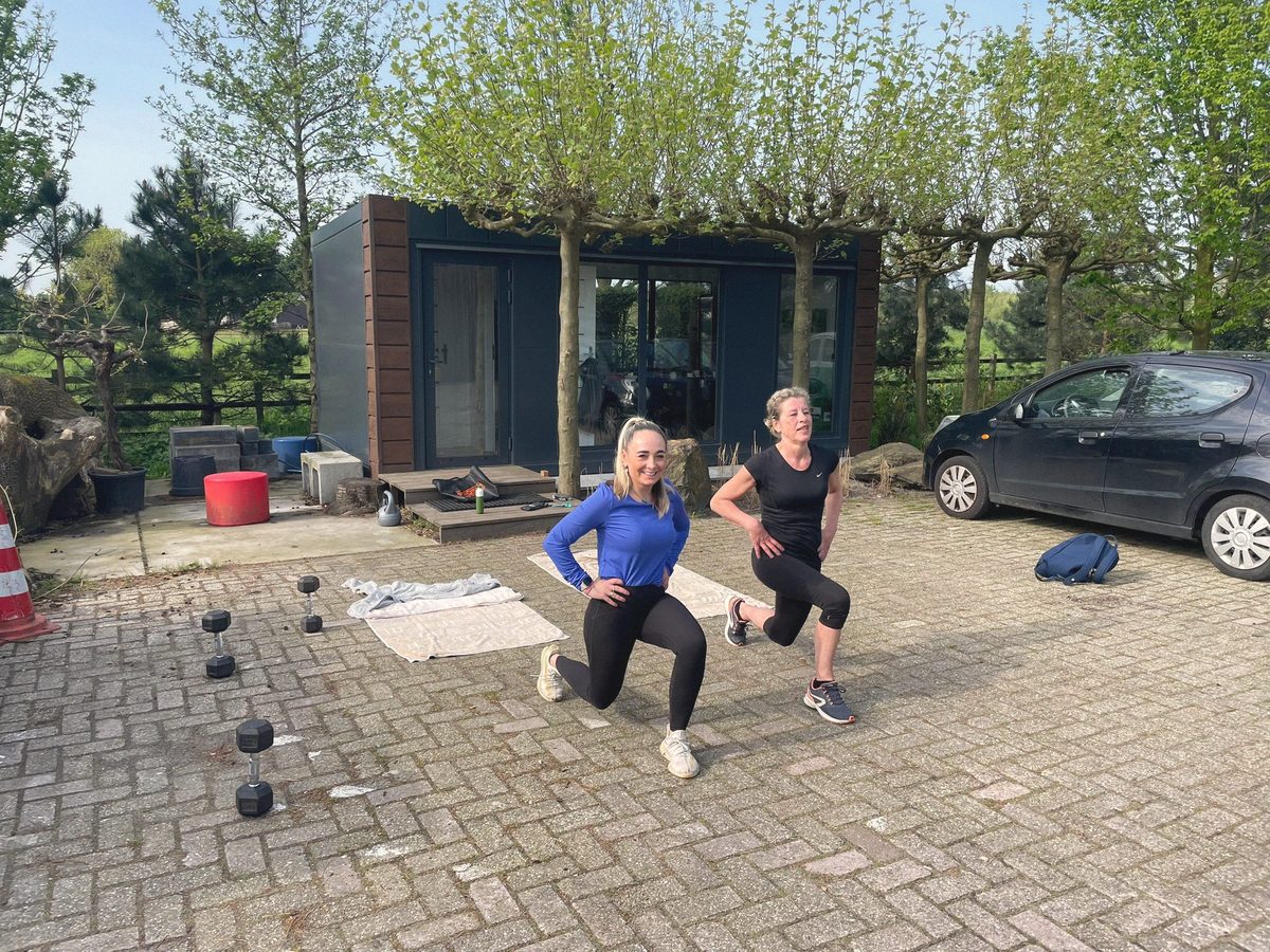 Twee mensen doen tegelijk een uitvalspas op een oprit, met kettlebells klaar